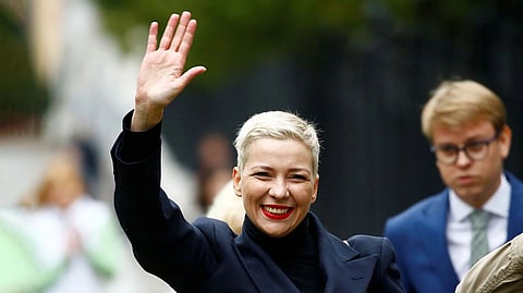 Former political prisoner Maria Kolesnikova with short blonde hair waves enthusiastically, wearing a black outfit. A blurred person in a blue suit is in the background.