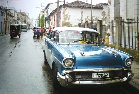 A street in Santa Clara, Cuba, a blue vintage car is parked, some people are gathered in the background