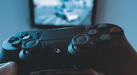 Close-up of a person holding a PlayStation controller with a blurred video game on screen in the background, conveying a focus on gaming.
