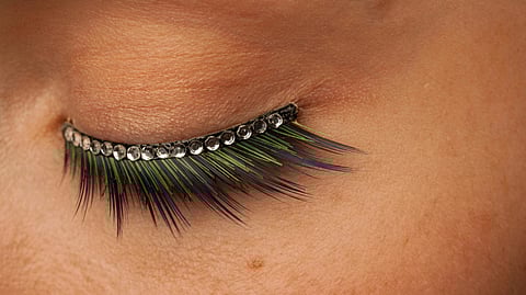 Close-up of a closed eye with long, colorful green and blue eyelashes, adorned with a row of small rhinestones, creating a sparkling effect.