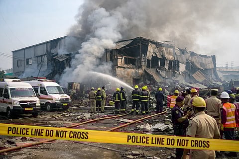 Firefighters battle a large industrial building fire, smoke billowing. Emergency crews and vehicles are on-site, with police tape in the foreground.