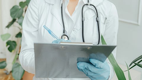 Smiling female doctor wearing protective eyewear holds a clipboard and pen. She's dressed in a white coat with a stethoscope, conveying professionalism.