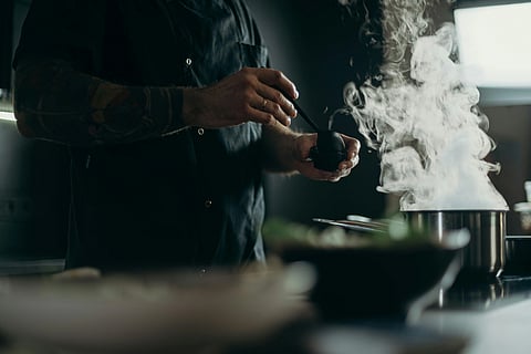 A chef, with tattooed arms, seasons a dish beside a steaming pot in a dimly lit kitchen, creating a warm and focused cooking atmosphere.