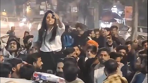 A group gathered in front of a police car, with a woman in white standing above thr crowd with an arm raised.
