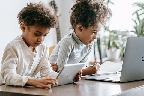 Two children focus intently on a tablet and laptop at a wooden table, with plants and a guitar in the background, conveying a studious atmosphere.