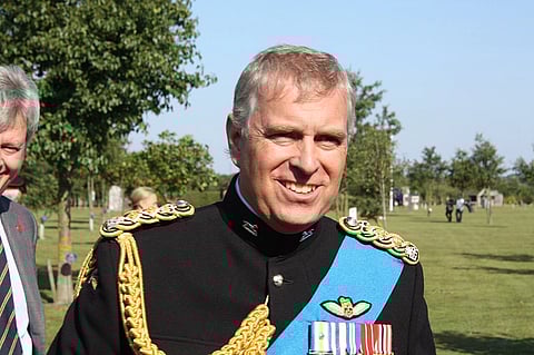 Andrew Mountbatten-Windsor in a decorated military uniform stands smiling in a green park under a clear blue sky. He wears a blue sash and medals, conveying formality.