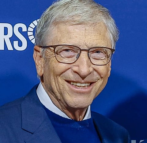 Portrait of Bill Gates  with glasses smiling, wearing a blue suit and sweater over a white shirt. Blue backdrop features partial white text and a logo.