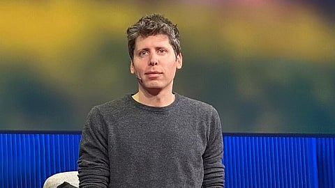 OpenAI CEO Sam Altman in a gray sweater and blue jeans sits on a chair against a colorful blurred background. They are wearing blue sneakers and hold a device, appearing thoughtful.