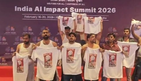 A group of men on stage at the India AI Impact Summit 2026 hold up white T-shirts with printed designs, appearing organized and determined.