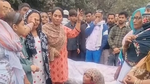 A group of people stand in a crowd around a body covered in a shroud.