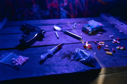 Dark, moody scene on a wooden table with scattered pills, syringe, small bags, spoon, cigarette, and handgun, conveying a theme of danger and illicit activities.