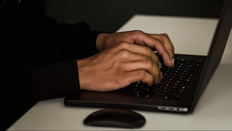 A person in a dark hoodie types intently on a laptop in a dimly lit room, conveying a sense of mystery or secrecy.
