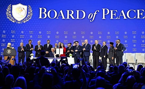 Image of Board of Peace Charter Announcement and Signing Ceremony January 2026.  A group of people stand on stage against a "Board of Peace" backdrop. A central figure sits holding a signed document, surrounded by applauding individuals. The atmosphere is formal and celebratory.