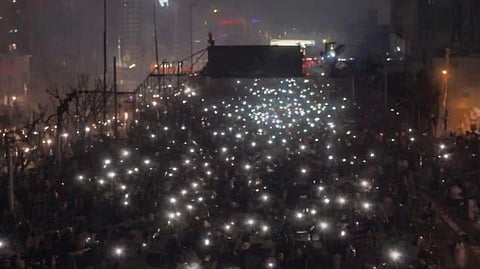 A large nighttime crowd holds up phone flashlights, creating a sea of white lights. The scene conveys unity and peaceful gathering in a dense urban setting.