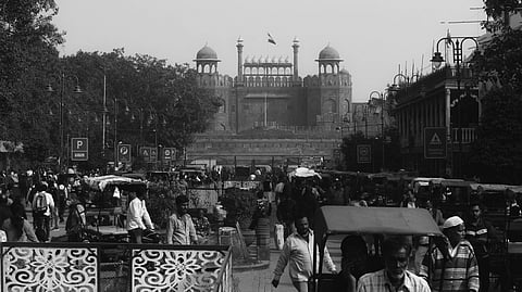 Black and white image of a bustling street leading to the historic Red Fort in Delhi. People, vehicles, and market stalls fill the vibrant scene.