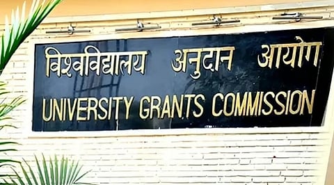 Black sign with gold lettering reads "University Grants Commission" in English and Hindi on a white brick wall, with green leaves visible on the left.