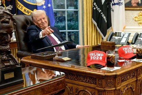 Image of Donald Trump in a suit sits confidently at a wooden desk, pointing. On the desk are red "Trump 2028" hats and glasses of soda. Flags and a statue are in the background.