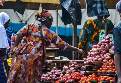 A vibrant market scene with women in colorful attire, selling fresh tomatoes, peppers, and onions displayed in baskets, conveying a lively, bustling atmosphere.