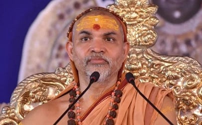 Image of Swamy Avimukteshwaranand  in orange robes sits on an ornate golden chair. He has a tilak on his forehead and holds a microphone.