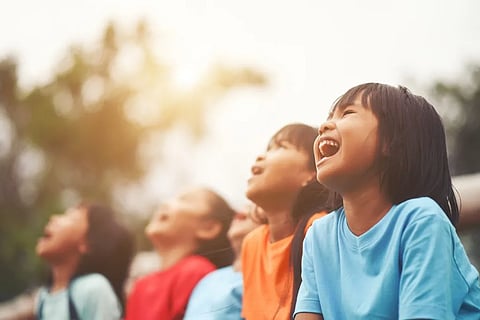 A group of children in colorful shirts laugh joyfully, gazing upwards in an outdoor setting. Sunlight filters gently through blurred trees in the background.