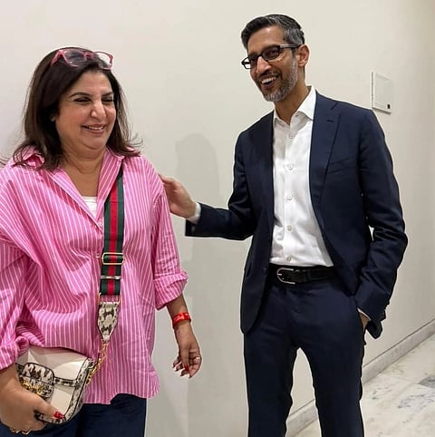 A woman in a pink striped shirt smiles next to a man in a dark suit. They are standing against a plain wall, engaged in a friendly conversation.