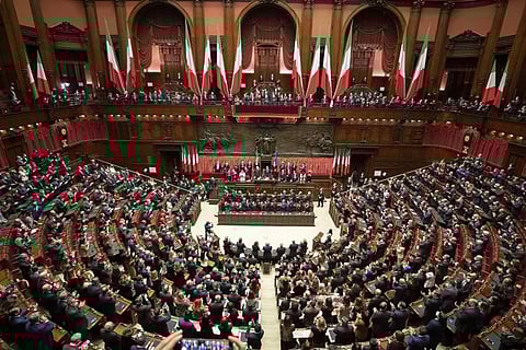 A vast, ornate parliamentary chamber filled with seated delegates. Italian flags hang prominently, creating a formal, official atmosphere.