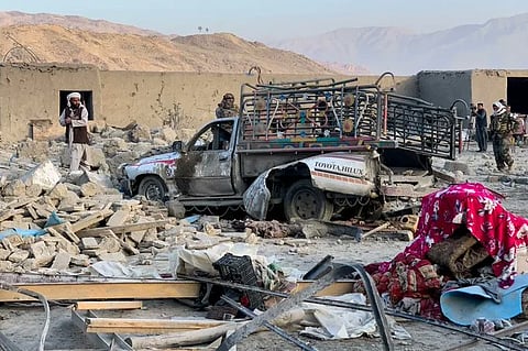 A damaged pickup truck amidst rubble from destroyed buildings in a barren landscape. People survey the scene, conveying a sense of devastation and loss.