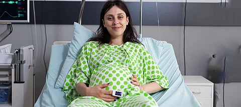 A woman in a green dotted gown lies while smiling on a hospital bed, holding her rounded belly.