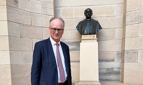 Matt Riddley with glasses stands beside a bronze bust of Sir Edwin Lutyens on a pedestal, set against a stone wall.