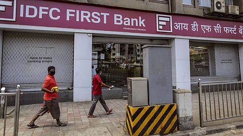 A branch of IDFC FIRST bank with two shutters down and two people in red walking in front.