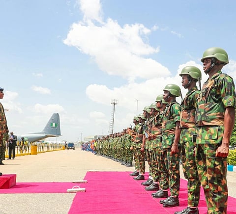 Soldiers in camouflage uniforms and helmets stand in formation on a pink carpet. A military officer inspects them. A plane is visible under a blue sky.