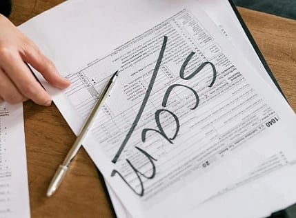 A person holding a pen over a tax form marked with "scam" in bold letters, conveying a warning about fraudulent documents.