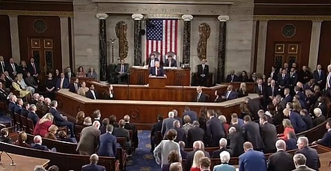 The image depicts a formal gathering in a oval office with officials seated and standing. President Trump stands at a podium before a U.S. flag. The scene conveys seriousness and attentiveness.