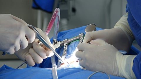 Surgical team performing surgery, hands in sterile gloves using medical instruments over a patient covered in blue drapes. Bright, clinical setting.
