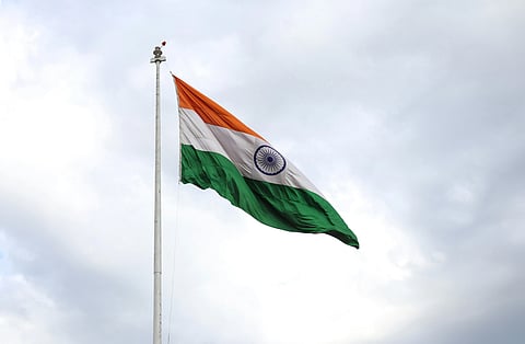 The Indian national flag waves against a cloudy sky. Its saffron, white, and green stripes with the blue Ashoka Chakra convey a sense of pride and patriotism.