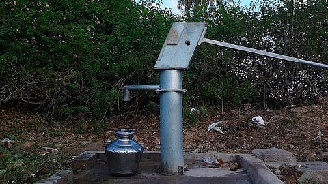 A groundwater pump with a lota under the tap and shrubs behind.