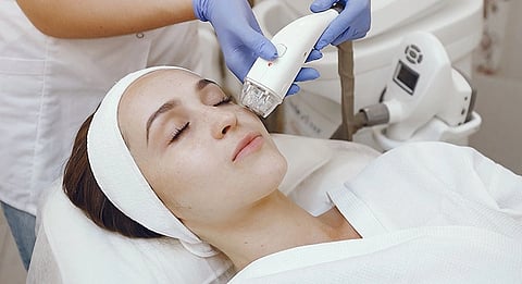 A woman receiving a facial treatment in a spa setting. She's wearing a headband and robe, eyes closed, while a beautician applies a device to her face.