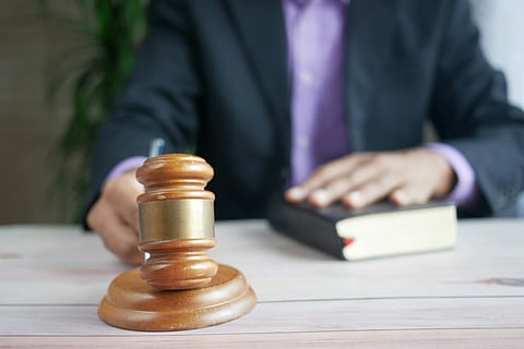A person in a suit, partially visible, holds a book while a wooden gavel is in the foreground on a table. The setting suggests a legal or judicial theme.