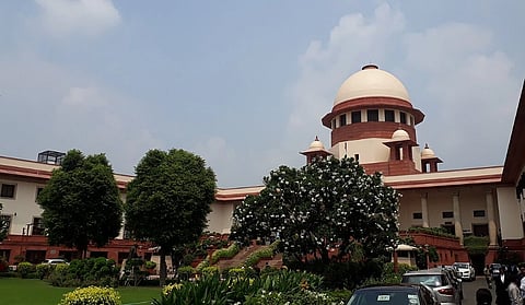 Image of Inside the Supreme Court of India, Bhagwandas Road, New Delhi. Exterior of a large courthouse with a prominent domed structure, surrounded by lush green gardens and trees under a partly cloudy sky. Cars are parked nearby.