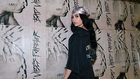 A woman in a balck t-shirt and bandana stands in front of a wall plastered with posters.