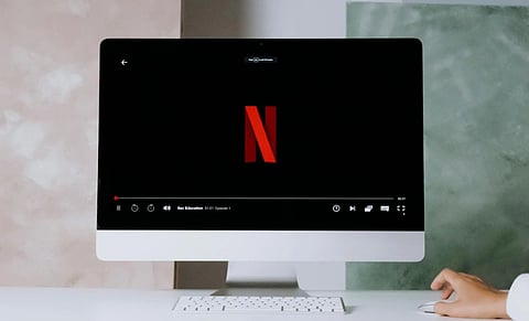 A computer screen displays the Netflix logo with a paused show. A hand reaches for a mouse on a white desk, conveying a sense of anticipation.