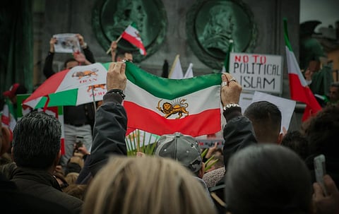 Crowd at a protest holds flags with lion and sun emblem; signs include "No to Political Islam." The mood is tense and the setting is urban.
