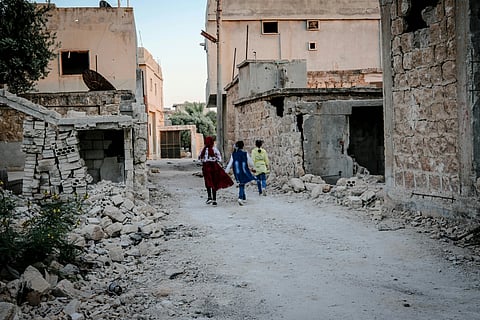In the image broken buildings are shown with blasts showing a war environment with three childrens walking in between these broken buildings