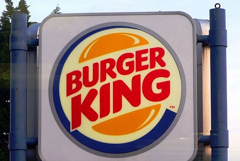 Burger King logo on a square sign, featuring bold red text inside a stylized burger with orange buns. Background is partly obscured by trees.