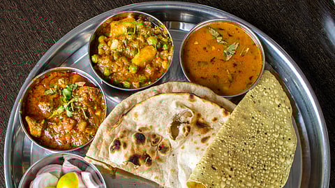 A stainless steel thali with colorful Indian dishes: two curries, dal, roti, papad, sliced onions, lemon, yellow rice, and a glass of yogurt.