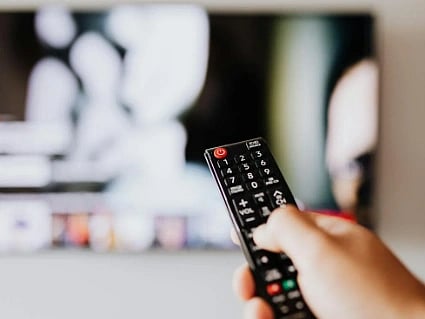 A hand holds a TV remote pointed at a blurred television screen. The scene suggests an intention to change channels or adjust volume.