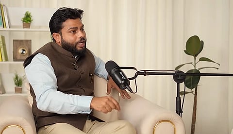 Raghav Trivedi in a brown vest speaks into a microphone during a podcast. He's seated on a light-colored sofa in a cozy, plant-decorated room. Engaged and expressive.