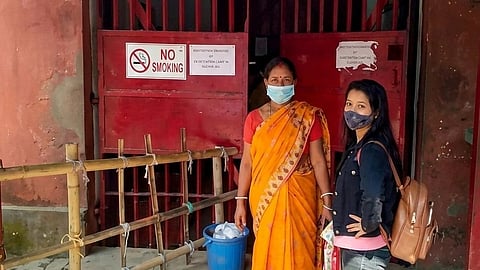 two women in masks stand in front of a red gate with a no-smoking sign.