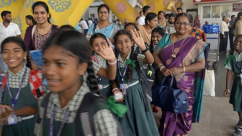 A group of smiling schoolchildren in green uniforms, waving and carrying backpacks, walk with teachers past a colorful, patterned structure indoors.