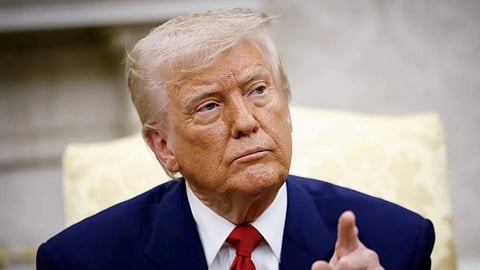 Image of Donald Trump with his classic blue suit and his red tie. His fingers are pointed towards upward. The background is blurred. 